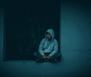 Perempuan muda duduk di tepi jendela saat hujan, menatap keluar dengan ekspresi reflektif.