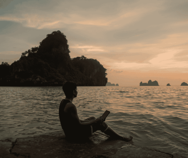 Pria duduk di tepi pantai saat senja sambil menatap laut dengan ekspresi rindu, berlatar nuansa timur Indonesia.