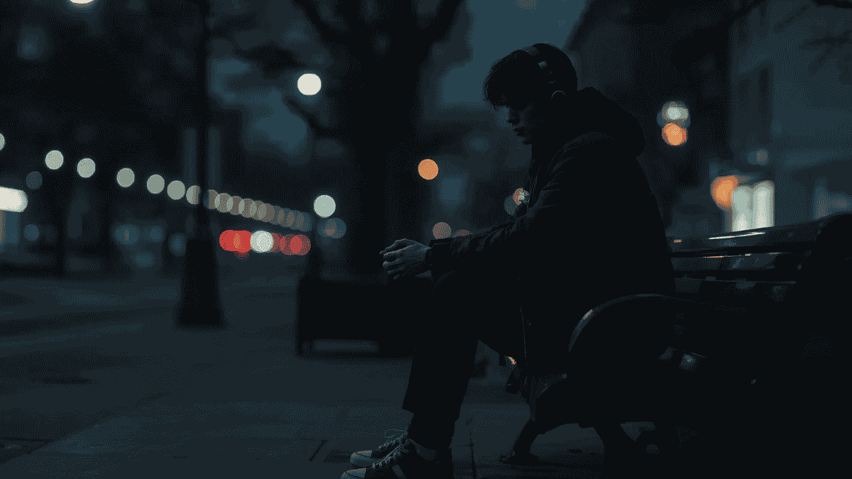 Anak muda termenung sendirian di bangku kota saat senja, mengenakan headphone.